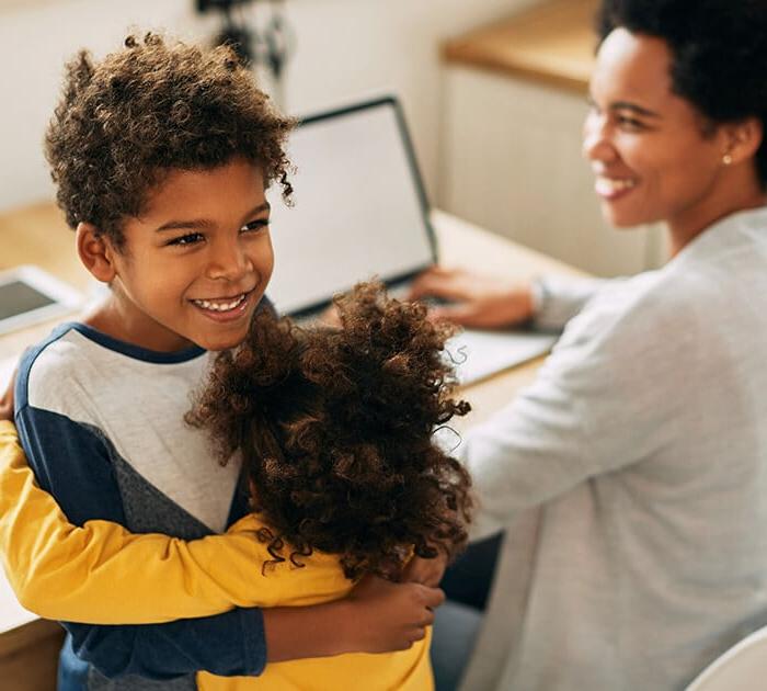 A brother and sister are hugging while their mom is on a laptop